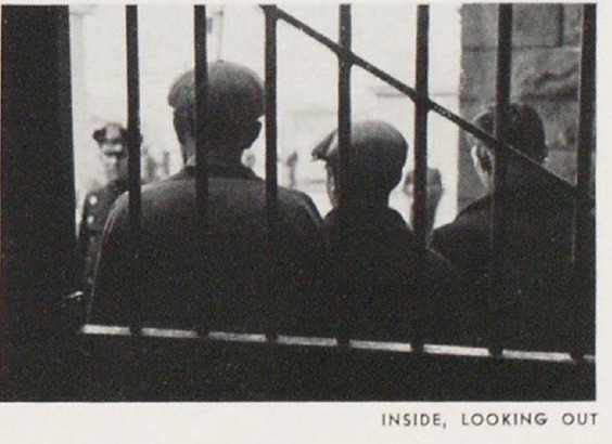 An image from Blackwell's Penitentiary, in the days before Rikers Island. Pictured are several men, wearing caps, standing behind bars. Image is black and white